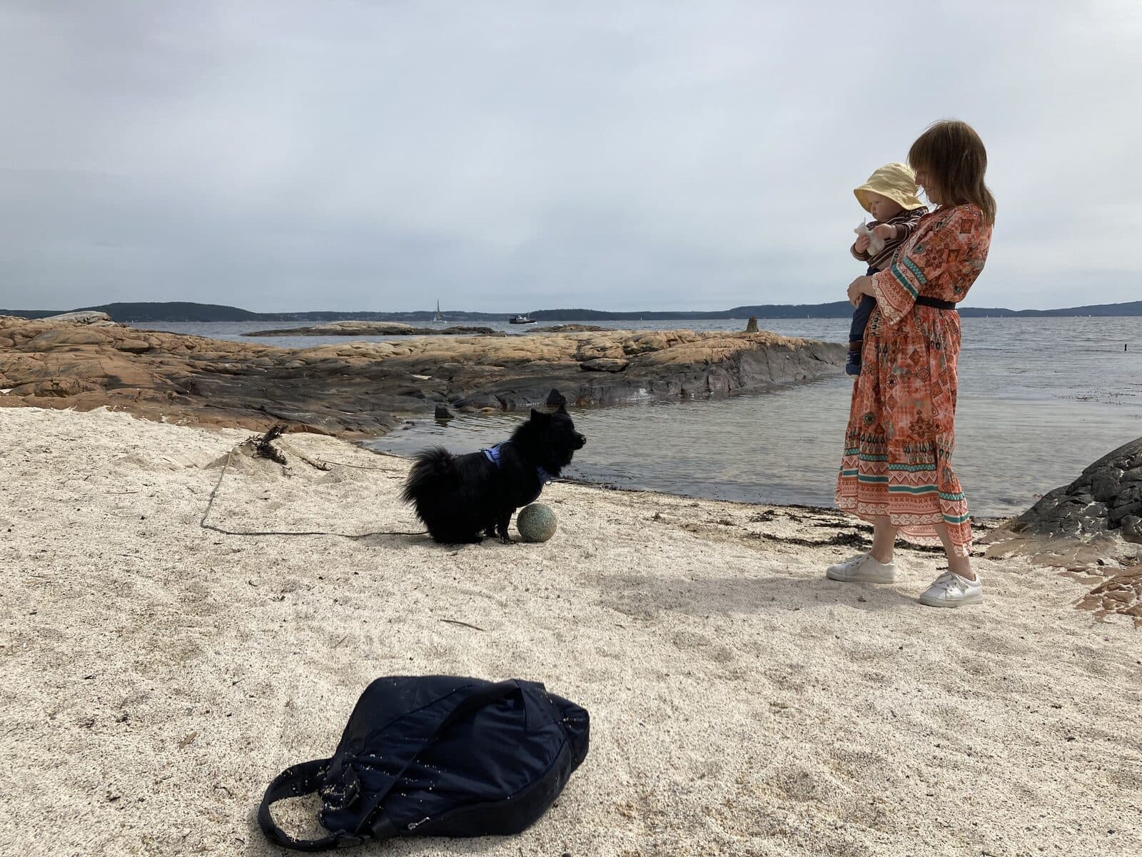 En mor med et barn på armen og en svart hund på en strand